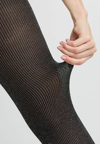 Collant neri a motivo con fili metallici argento, caratterizzati da una texture a costine e un tessuto liscio e elastico. Mano che tiene il tessuto.