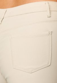 Gros plan sur un tissu beige de pantalon ajusté, avec une poche arrière unique à couture contrastée et une texture lisse.