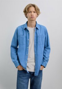 Camisa de mezclilla azul claro con un diseño de botones, dos bolsillos frontales y mangas largas. Llevada sobre una camiseta blanca y combinada con jeans azules.
