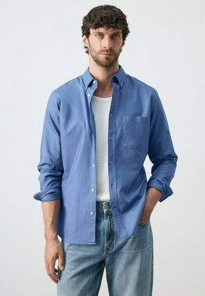 Hombre con cabello rizado oscuro, vistiendo una camisa azul de botones sobre una camiseta blanca y jeans azul claro, de pie con una mano en el bolsillo.