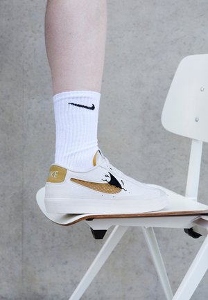 Stopa w białym bucie sportowym Nike ze złotymi i czarnymi akcentami spoczywająca na białym krześle na szarym tle.