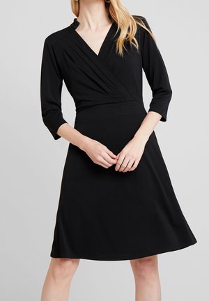 Mujer con vestido negro hasta la rodilla, mangas tres cuartos y cuerpo plisado con escote en V, manos entrelazadas a la altura de la cintura.