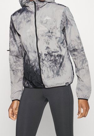 Personne portant une veste à capuche zippée tie-dye grise et noire, avec des leggings noirs, debout devant un fond clair uni.