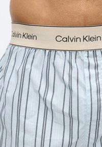 Λεπτομέρεια μέσης ριγωτών ανοιχτομπλε και γκρι μποξεράκια Calvin Klein με μπεζ ελαστική μέση που φέρει μαύρο λογότυπο.