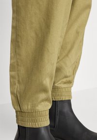 Olivgröna byxor med elastiska mudd, tillverkade av mjukt tyg, tillsammans med svarta ankelskor, med en slank design och minimal detaljering.