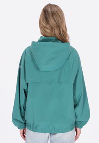 Kurtka z kapturem w kolorze morskim o luźnym kroju, z elastycznymi mankietami i wentylowanym tyłem. Gładka faktura z minimalistycznymi detalami, zestawiona z jasnoniebieskimi dżinsami.