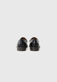 Mocasines de cuero negro con una textura suave, puntera redondeada y tacón bajo marrón. Presentan detalles de costura sutil a lo largo de las costuras.