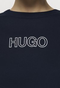 Primo piano di una persona che indossa una maglietta a girocollo blu navy con il testo bianco contornato "HUGO" centrato sul petto.