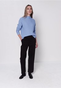 Pull en maille côtelée bleu clair avec col montant, associé à un pantalon noir sur mesure et des chaussures noires, le tout sur un fond neutre.