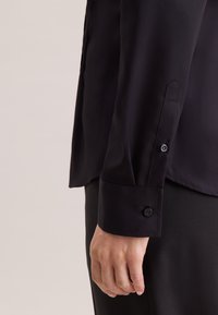 Zbliżenie na ramię osoby w czarnej koszuli z długim rękawem z mankietami na guziki na neutralnym tle.