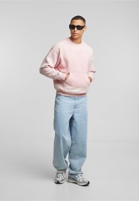 Felpa rosa chiaro con collo rotondo, tasca frontale e logo bianco. Abbinata a jeans baggy azzurri e scarpe da ginnastica argentate.