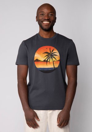 Donkergrijze t-shirt met een ronde afbeelding van een zonsondergang met een palmboom en bergen, met levendige oranje, gele en blauwe tinten.