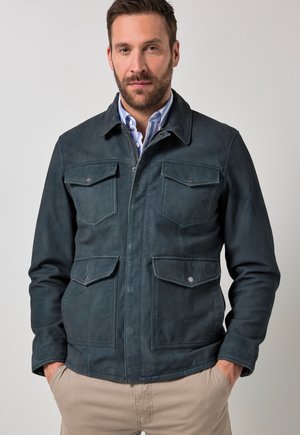 Hombre con chaqueta azul oscuro con cuatro bolsillos con botones, camisa de rayas azules, pantalones beige, de pie con las manos en los bolsillos contra un fondo liso.