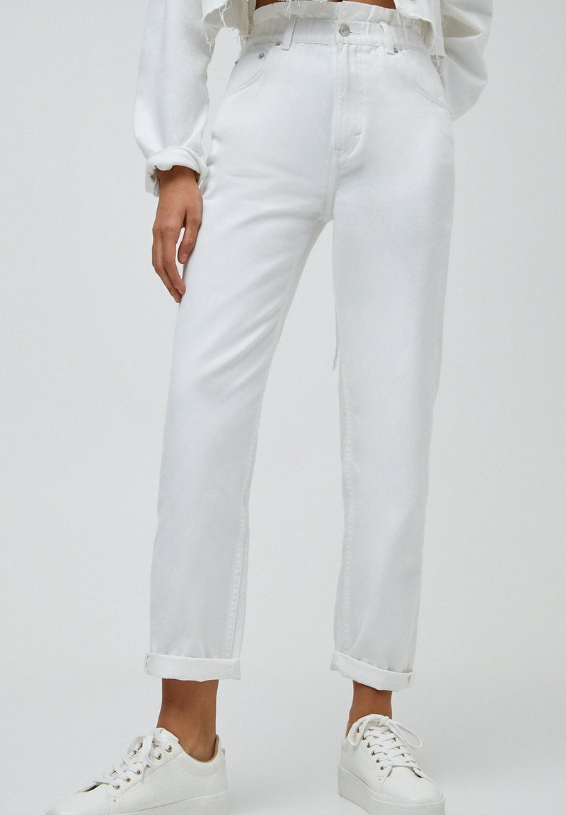 Personne portant un jean blanc taille haute avec les revers roulés et des baskets blanches, debout devant un fond clair uni.