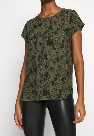 Vrouw draagt een olijfgroen kortärmlig top met zwarte tijgerprint en zwarte neplederen legging, tegen een effen achtergrond.