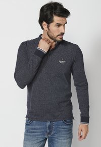 Mornarski polo majica s dugim rukavima od teksturirane tkanine, s kontrastnim ovratnikom i manžetama, te malim logotipom na prsima. Nosena s plavim trapericama.