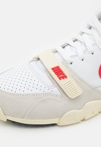 Witte sneaker met grijze leren panelen en perforaties. Bevat rode "Nike" branding en een beige band. Rubberen zool met gestructureerde accenten.