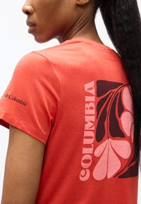 Κοντινό πλάνο ενός ατόμου που φοράει κόκκινο μπλουζάκι Columbia με μεγάλο λουλουδάτο σχέδιο και το όνομα της μάρκας στην πλάτη και στο μανίκι.