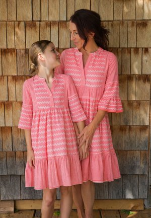 Mother and daughter wearing matching pink patterned dresses, holding hands and looking at each other in front of a wooden shingle wall.
