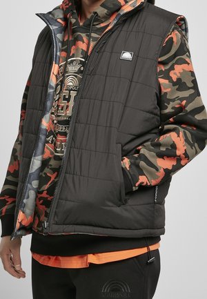 Personne portant une veste matelassée noire par-dessus un sweat à capuche camouflage orange, vert et noir avec un motif graphique et un pantalon noir avec logo.
