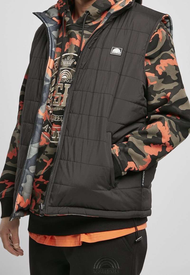 Personne portant une veste matelassée noire par-dessus un sweat à capuche camouflage orange, vert et noir avec un motif graphique et un pantalon noir avec logo.