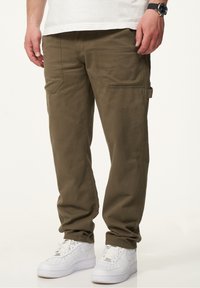 Olivgrüne Cargo-Hosen mit mehreren Taschen, einer lockeren Passform und einer glatten Textur, kombiniert mit weißen Sneakers.