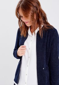 Veste texturée bleu marine avec fermeture éclair à l'avant et poches latérales, portée sur une chemise blanche à col. Le tissu semble doux et structuré.