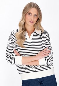 Maglione a righe in bianco e nero, con colletto a V e polsini a coste, mostrato con le braccia incrociate su uno sfondo bianco.