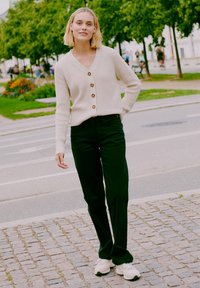 Beige pletený cardigan s dřevěnými knoflíky, v kombinaci s tmavozelenými kalhotami a bílými teniskami, zasazený do zeleného venkovního prostředí.