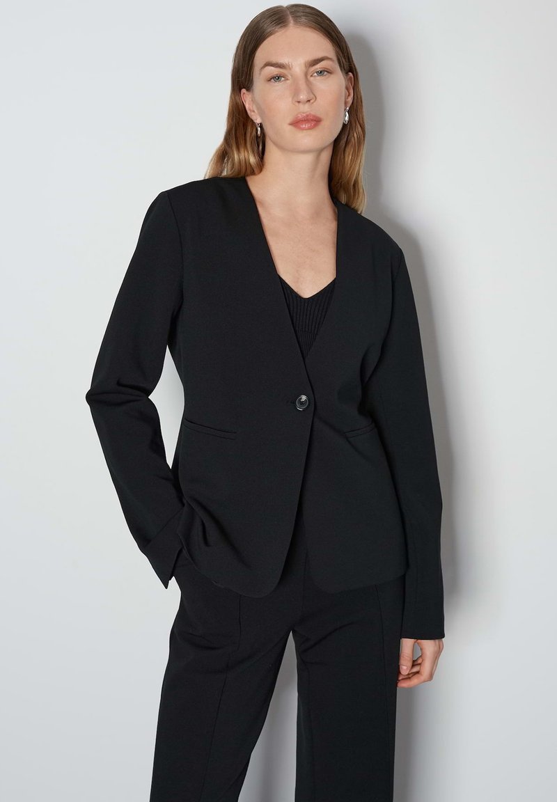Blazer noir ajusté avec un design épuré, fermeture à un bouton et revers crantés. Tissu lisse avec poches latérales.