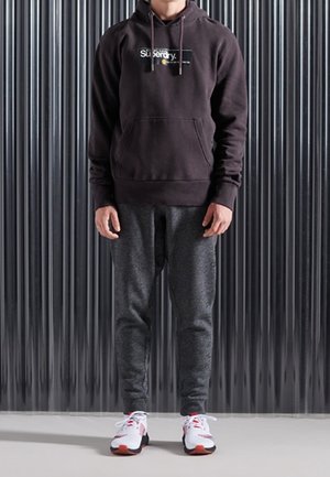 Osoba ubrana w czarną bluzę Superdry z kapturem, ciemnoszare spodnie dresowe oraz białe sneakersy z czerwonymi akcentami, stojąca na tle czarnej falistej ściany.