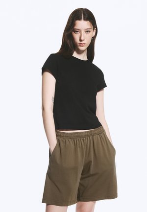 Jeune femme aux longs cheveux foncés portant un t-shirt noir à manches courtes et un short marron ample, debout les mains dans les poches sur un fond blanc.
