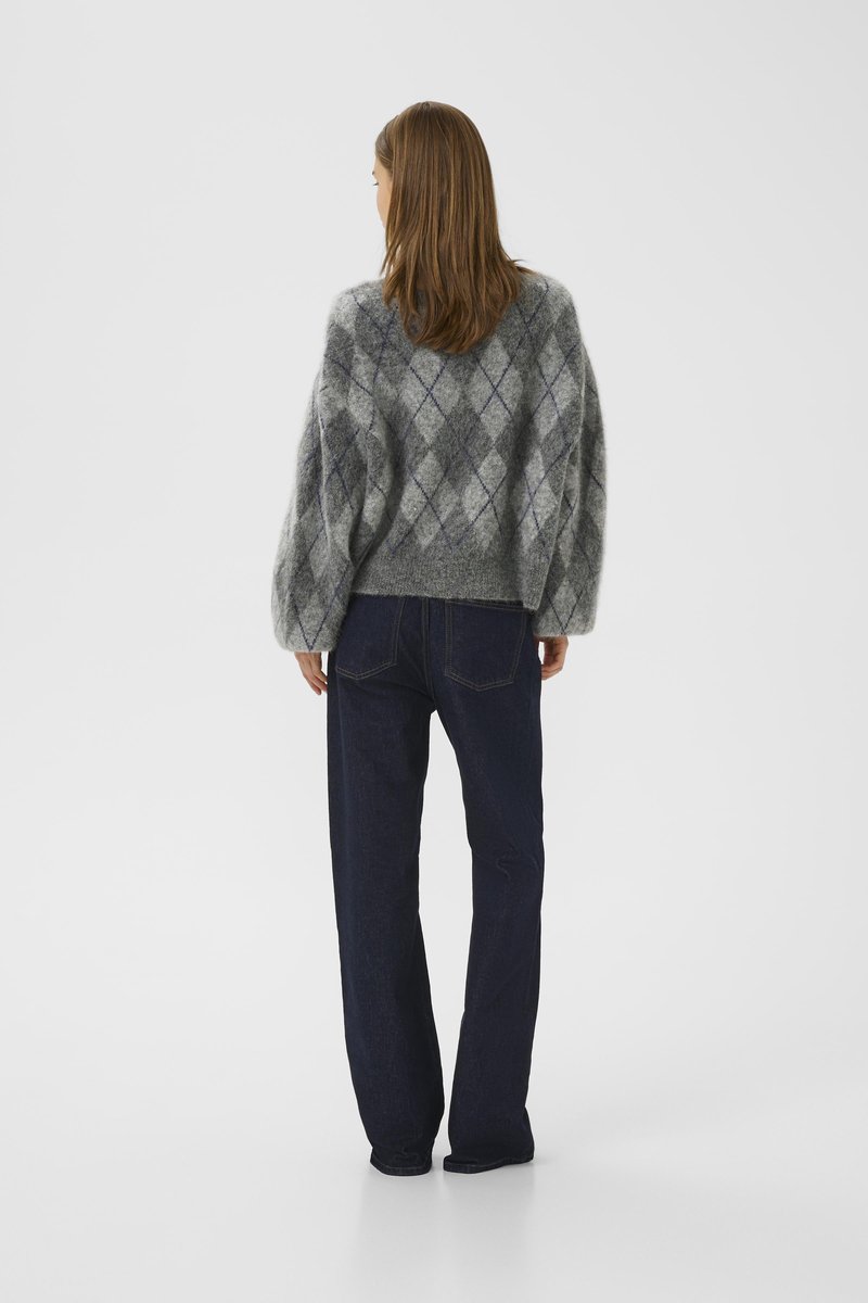 Kvinna som står vänd bort, iförd en grå rutig tröja och mörkblå raka jeans på en enfärgad ljus bakgrund.