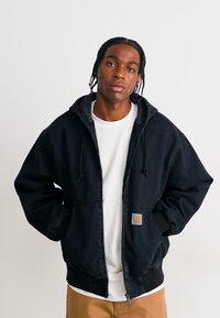 Young man with braided hair wearing a black zip-up hoodie over a white shirt and tan pants, standing with hands in pockets against a plain background.