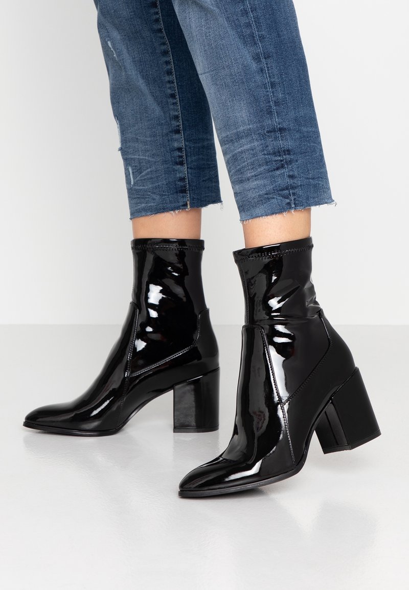 Bottes noires en cuir verni à cheville avec un bout pointu, un talon épais, un design épuré et des détails élastiques, portées avec un jean court en denim.