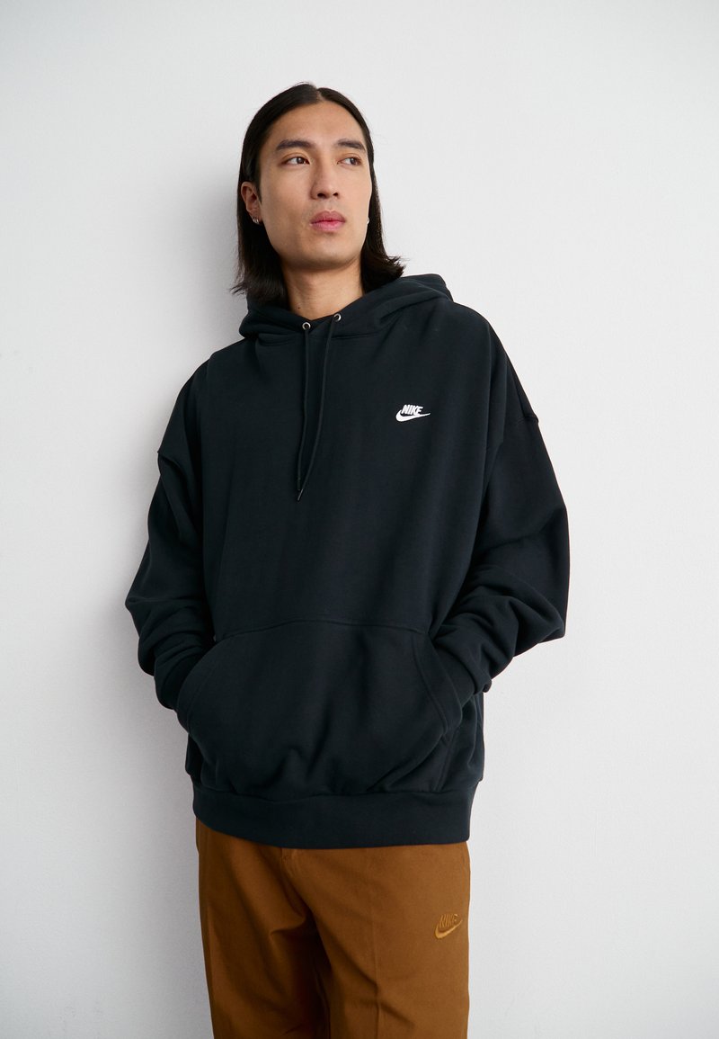 Sweatshirt à capuche noire avec une poche avant, arborant un logo Nike blanc sur la poitrine. Associé à un pantalon marron. Tissu lisse et décontracté.