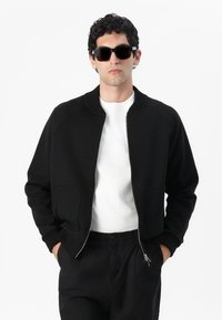 Chaqueta bomber negra con cremallera frontal, cuello acanalado y dos bolsillos laterales, combinada con una camiseta de cuello redondo blanca y pantalones oscuros.