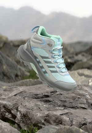 Une chaussure de randonnée menthe et grise avec une semelle épaisse flotte au-dessus d'un terrain rocheux, avec des montagnes brumeuses en arrière-plan.