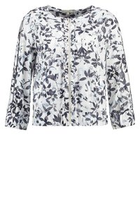 Veste blanche avec imprimé floral noir et gris, fermeture éclair devant, manches longues, et léger détail volanté le long de la ligne de la fermeture éclair.