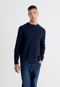 Tumši zils adīts džemperis ar teksturētu rakstu, apaļu kakla izgriezumu un ribota aproce un apakša. Apvienots ar ziliem džinsa biksēm.