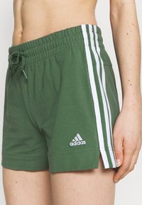 Primer plano de una persona que lleva pantalones cortos verdes de Adidas con rayas blancas y cintura con cordón, mostrando el abdomen y la mano derecha.