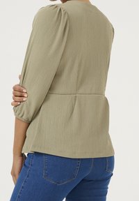 Person iført en strukturert olivengrøn puffærmet bluse og blå denimjeans, vist fra ryggen med den ene arm bøjet over kroppen.