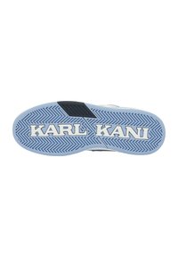 Semelle de sneaker blanche avec des accents bleus, présentant un motif de bande de roulement en zigzag et le logo "KARL KANI" bien en vue. Surface texturée pour une meilleure adhérence.