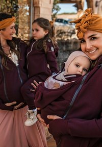 Due donne indossano giacche borgogna con zip, una delle quali tiene un bambino in una tasca frontale, mentre l'altra tiene una bambina. Sono visibili morbidi tessuti e dettagli di design pratici.
