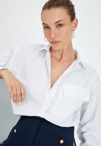 Camisa blanca de botones con un bolsillo en el lado izquierdo, fabricada en material ligero, con cuello clásico y corte holgado, combinada con pantalones de cintura alta en color navy y botones dorados.