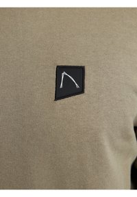 Tessuto di camicia khaki con texture a coste, caratterizzato da una toppa rettangolare nera con un design a linee bianche che rappresenta un arco, situata sul lato sinistro.