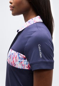 Γυναίκα που φοράει μπλε πουκάμισο πόλο με πολύχρωμο λουλουδάτο σχέδιο στο στήθος και το γιακά, με λογότυπο Calvin Klein στο μανίκι.