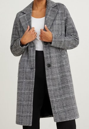 Personne portant un long manteau à carreaux gris et noir par-dessus un haut blanc et un pantalon noir, tenant les revers du manteau avec les deux mains.