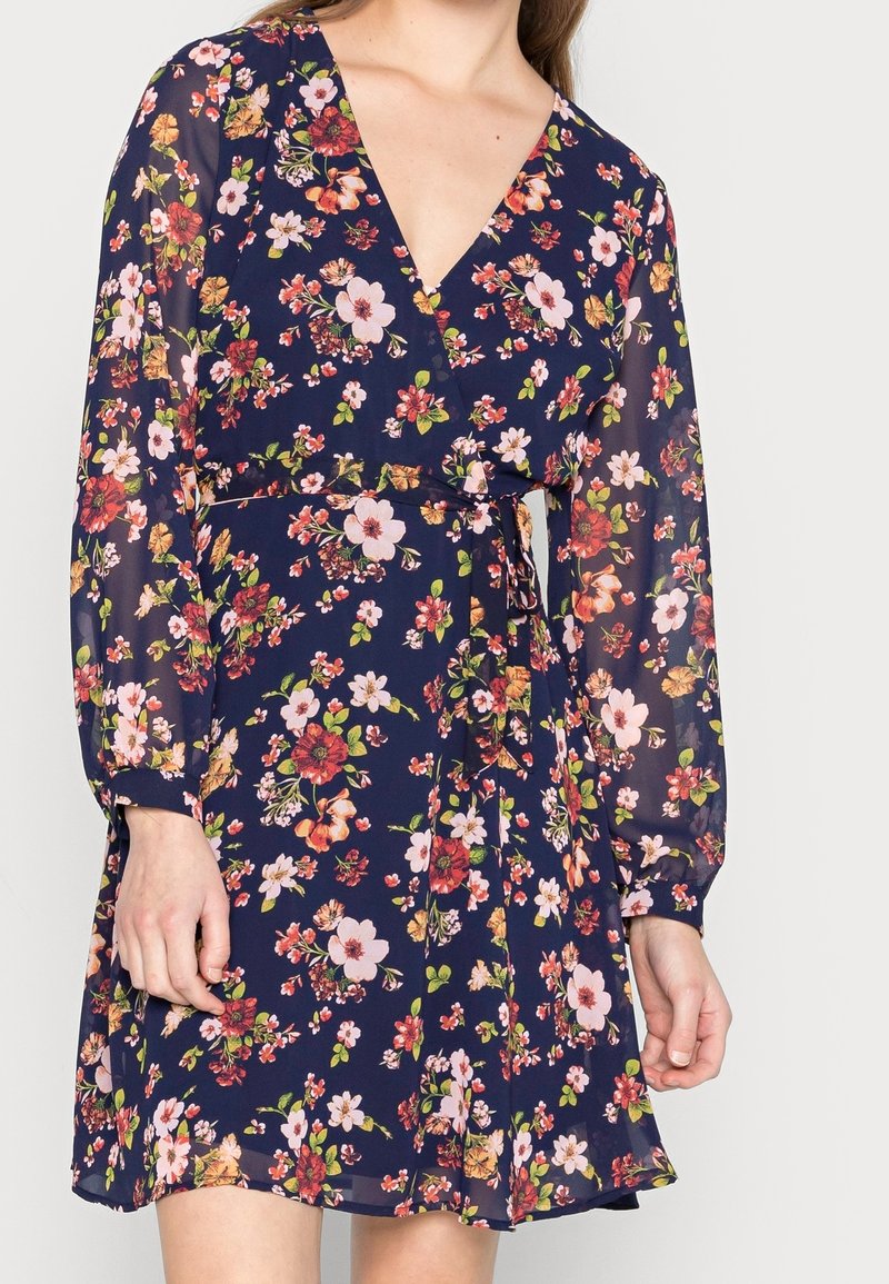 Floral wrap dress in navy, featuring long puff sleeves, a deep V-neck, and a tie waist. Textured fabric with multicolored flower patterns.