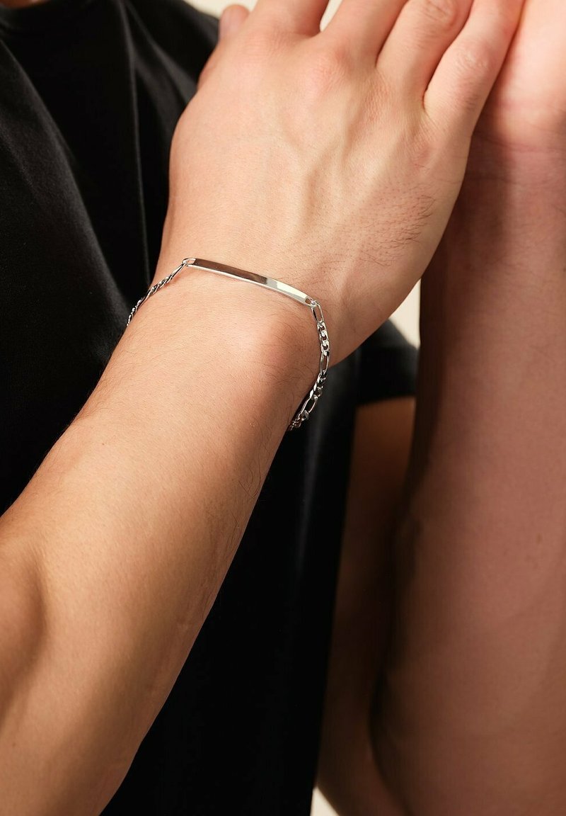 Bracciale d'argento con una elegante piastra rettangolare e un design a catena, indossato attorno a un polso, che mette in mostra una superficie metallica liscia.
